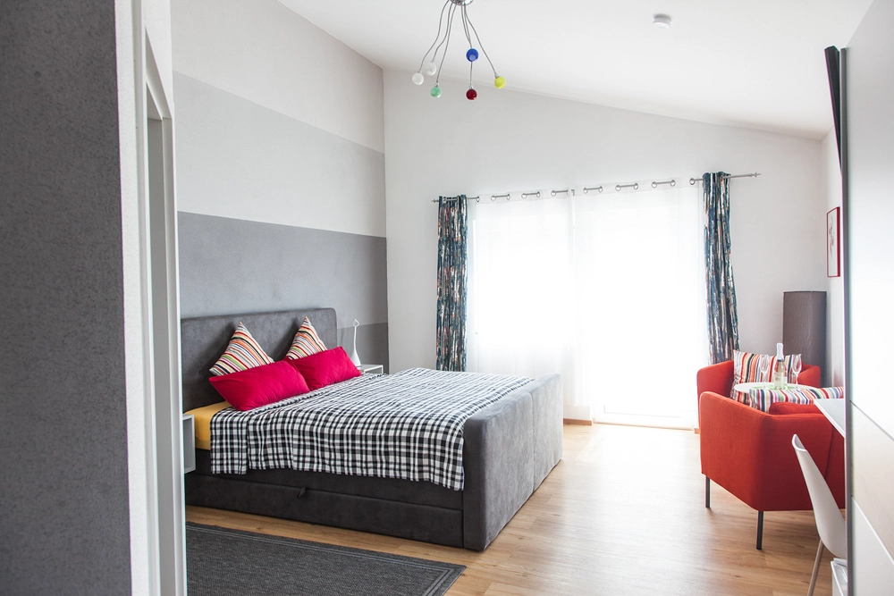 „Hahns Herberge“ – Schlafzimmer Innenansicht; Einrichtung in rot und grau gehalten, Lichtführung betont die Raumtiefe.